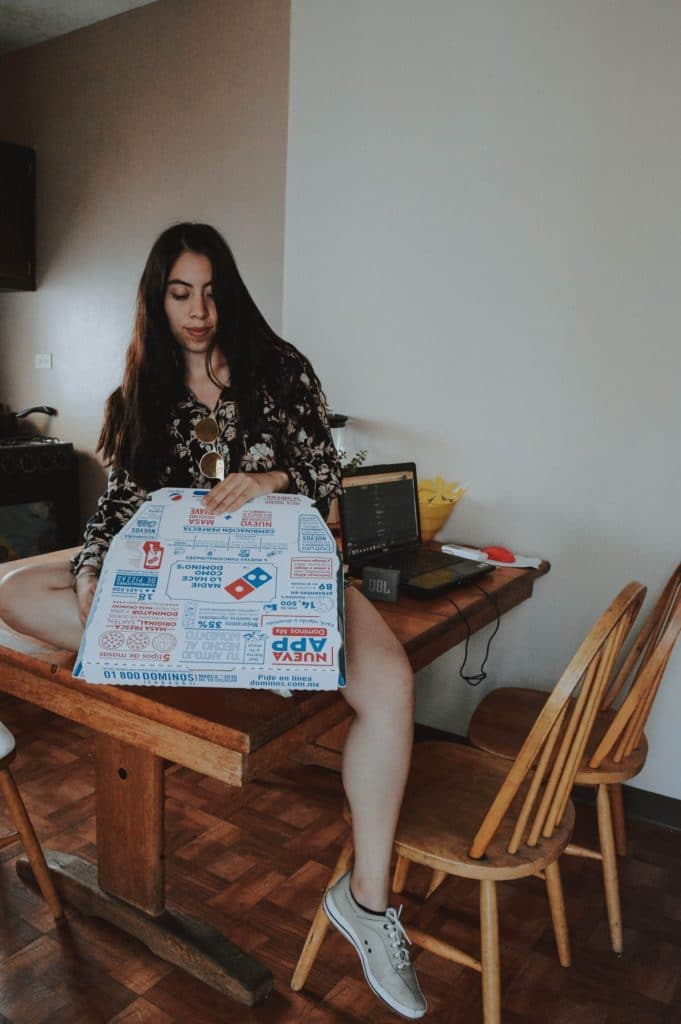 Female is sitting on a table top holding a box of cold Dominos pizza in the takeout box.